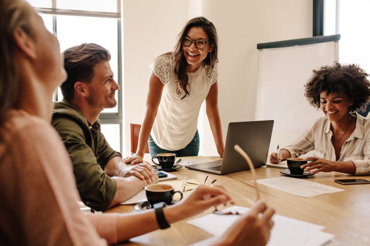 En gruppe på fire personer deltar i et møte hvor de diskuterer løsningen via purservice sin servicedesk. En kvinne med briller, som ser ut til å lede samtalen, står over bordet med en laptop foran seg, smilende og engasjert. To andre kvinner og en mann sitter rundt bordet, smilende mens de jobber med å løse ulike kundehenvendelser. Kaffekopper og notatblokker ligger på bordet, mens teamet samarbeider om å løse tickets knyttet til servicedesk-forespørsler.